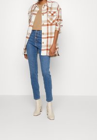 Camisa de cuadros en blanco, beige y naranja, superpuesta sobre una blusa beige, combinada con jeans azules y botas de tacón crema.