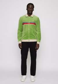 Zöld velúr zipzáras dzseki kontrasztos piros és fekete vízszintes csíkokkal, bézs gallérral, elülső zsebekkel, fekete nadrággal és fehér sportcipővel párosítva.