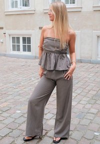 Femme blonde portant un top peplum à carreaux sans bretelles et un pantalon large assorti, debout dans une rue pavée près d'un bâtiment beige.