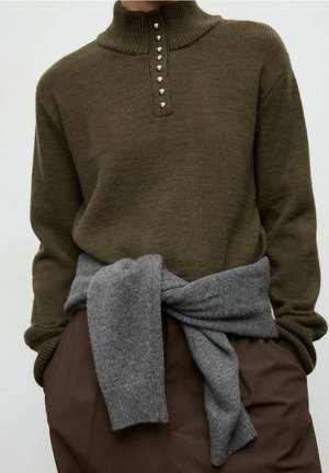 Pull en maille marron avec col boutonné porté sur un pantalon marron, pull gris noué autour de la taille, mains dans les poches.