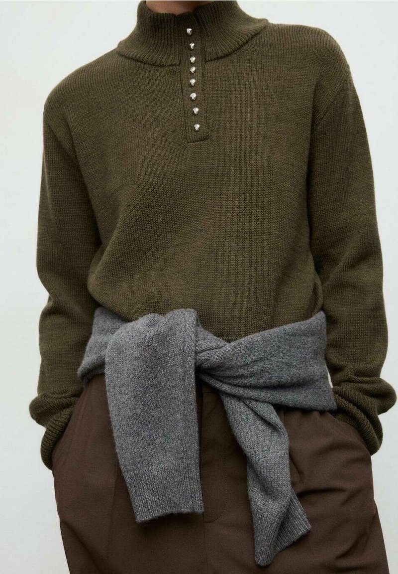 Pull en maille marron avec col boutonné porté sur un pantalon marron, pull gris noué autour de la taille, mains dans les poches.