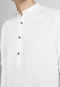 Vit långärmad skjorta med rund halsringning, med en knappslå som har tre bruna knappar och ett slätt, strukturerat tyg.