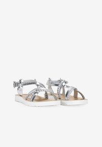 Sandalias plateadas con diseño entrelazado, acentos trenzados y una correa en el tobillo. Suela blanca y plantilla marrón para contraste. Estilo plano y abierto en la parte delantera.