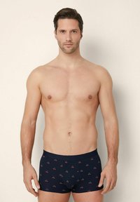 Uomo senza maglietta con capelli scuri corti, che indossa boxer blu navy con un motivo di piccole ciliegie rosse, in piedi su uno sfondo chiaro e semplice.