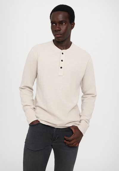 Lys beige langermet skjorte med henley-hals og tre sorte knapper. Brukt med mørkegrå tettsittende jeans. Glatt tekstur.