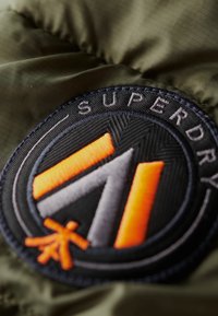 Parche circular negro con el texto "Superdry" y diseños geométricos en negrita en naranja y gris, sobre un fondo de tela acolchada verde oliva.