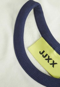 Indumento bianco con colletto a coste blu navy. Etichetta gialla brillante con testo nero che riporta "JJXX." Texture morbida, con un'enfasi sul design del colletto.