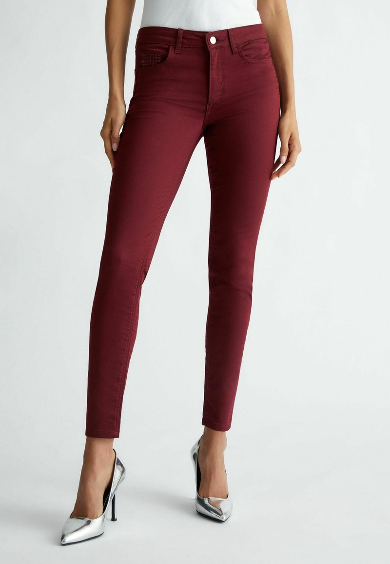 Femme portant un pantalon bordeaux ajusté et des talons hauts argentés, debout devant un fond clair uni.