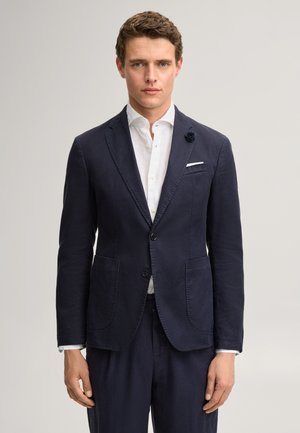 Mann trägt marineblauen Blazer mit zwei Knöpfen, weißem Hemd und passender marineblauer Hose, steht vor neutralem Hintergrund.