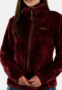 Giacca in pile color bordeaux con motivo a rombi, cerniera completa e colletto alto. Dettaglio in pelle marrone con logo vicino alla cerniera.