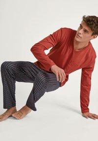 Rode blouse met lange mouwen en V-hals, voorzien van een zak, gecombineerd met gestreepte broek met afwisselend blauwe strepen op een donkere achtergrond.