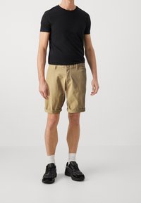 Černé tričko s kulatým výstřihem v kombinaci s béžovými šortkami s ohrnutím. Model má na sobě černé tenisky s bílými ponožkami, čímž vytváří neformální a přiléhavý vzhled.