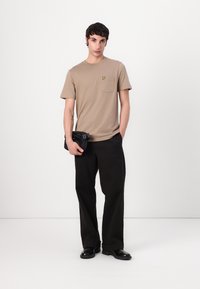 Beige bomull t-skjorte med lomme og en liten gul logo, kombinert med løse sorte bukser og sorte sko, holdt med en svart veske.