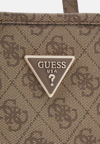 Sac à main en textile marron avec un logo en métal triangulaire portant "GUESS USA" et un design de point d'interrogation, motif texturé sur l'ensemble.