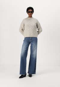 Lys grå strikket genser med et teksturert mønster, kombinert med vide blå denimjeans og svarte støvler, brukt med mørke solbriller.