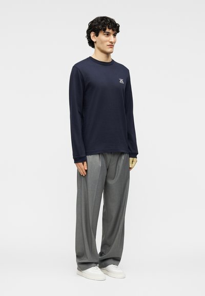 Άνδρας που στέκεται φορώντας μπλε-σκούρο μακρυμάνικο πουκάμισο, γκρι παντελόνια φαρδιάς γραμμής και λευκά αθλητικά παπούτσια, μπροστά σε απλό ανοιχτό φόντο.