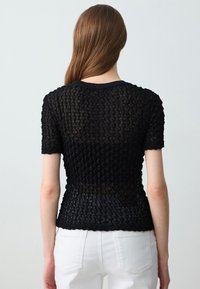 Donna con capelli castani lunghi che indossa un top nero a maniche corte con texture e pantaloni bianchi, vista di spalle su uno sfondo neutro.