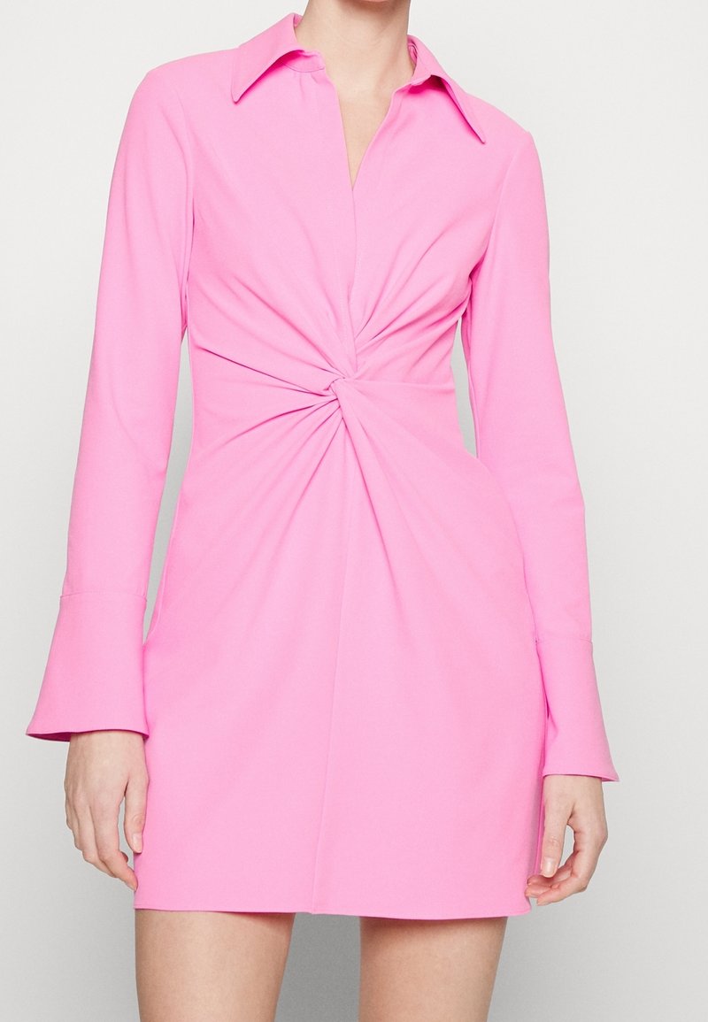 Robe rose à manches longues avec un col, dotée d'un détail noué à la taille et de manches évasées. Tissu lisse et extensible.