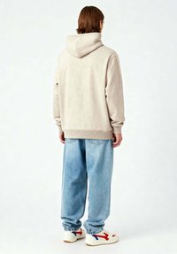 Felpa beige con cappuccio e vestibilità comoda, abbinata a jeans blu chiaro larghi e sneakers bianche con dettagli in rosso e blu.