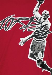 Camiseta roja con un gráfico en blanco y negro de una figura saltando y texto en negrita. El diseño tiene un aspecto texturizado con colores contrastantes.