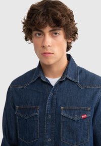 Camisa de denim en azul oscuro con dos bolsillos en el pecho, costuras claras y una pequeña etiqueta de logo roja. Presenta un cuello tipo polo y un diseño de botones en la parte delantera.