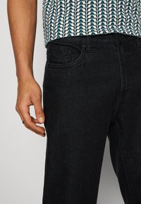Jean en denim noir avec un design classique, une coupe cinq poches et une texture lisse. Une main repose sur la ceinture.