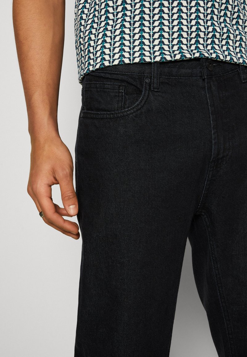 Schwarze Jeans aus Denim mit klassischem Design, fünf Taschen-Stil und glatter Textur. Eine Hand liegt auf dem Bund.