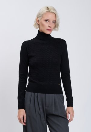 Femme aux cheveux courts blonds portant un pull à col roulé noir et un pantalon gris ample, debout devant un fond uni clair.