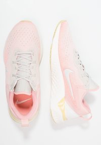 Un par de zapatillas de running Nike en rosa y beige, una vista desde arriba y la otra de lado, sobre un fondo blanco.