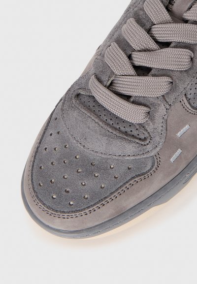 Punta di sneaker grigia in suede con perforazioni, lacci spessi coordinati e doppia cucitura su uno sfondo chiaro.