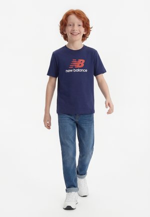 Băiat cu păr roșcat zâmbitor care merge, purtând un tricou bleumarin New Balance, blugi albaștri cu tiv și adidași albi pe fundal alb.