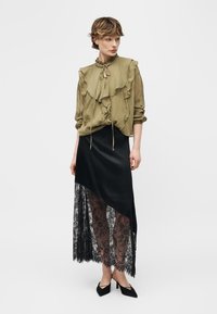 Personne portant un chemisier vert olive à volants avec un col noué, une jupe asymétrique noire avec une superposition de dentelle, et des chaussures à talons noires, debout devant un fond blanc.