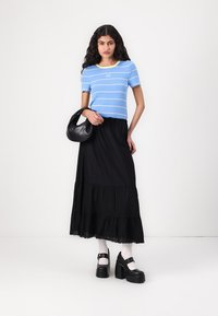 Top a maniche corte blu e bianche a righe con accenti gialli, abbinato a una gonna maxi nera a strati. La modella tiene una borsa nera e indossa scarpe con zeppa.
