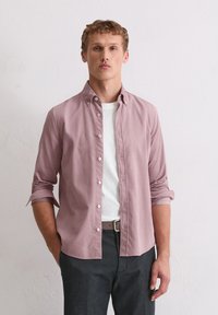 Jasnoróżowa koszula zapinana na guziki w luźnym kroju, z podwiniętymi rękawami, w zestawie z białym t-shirtem z okrągłym dekoltem i ciemnymi spodniami. Gładka tekstura materiału.