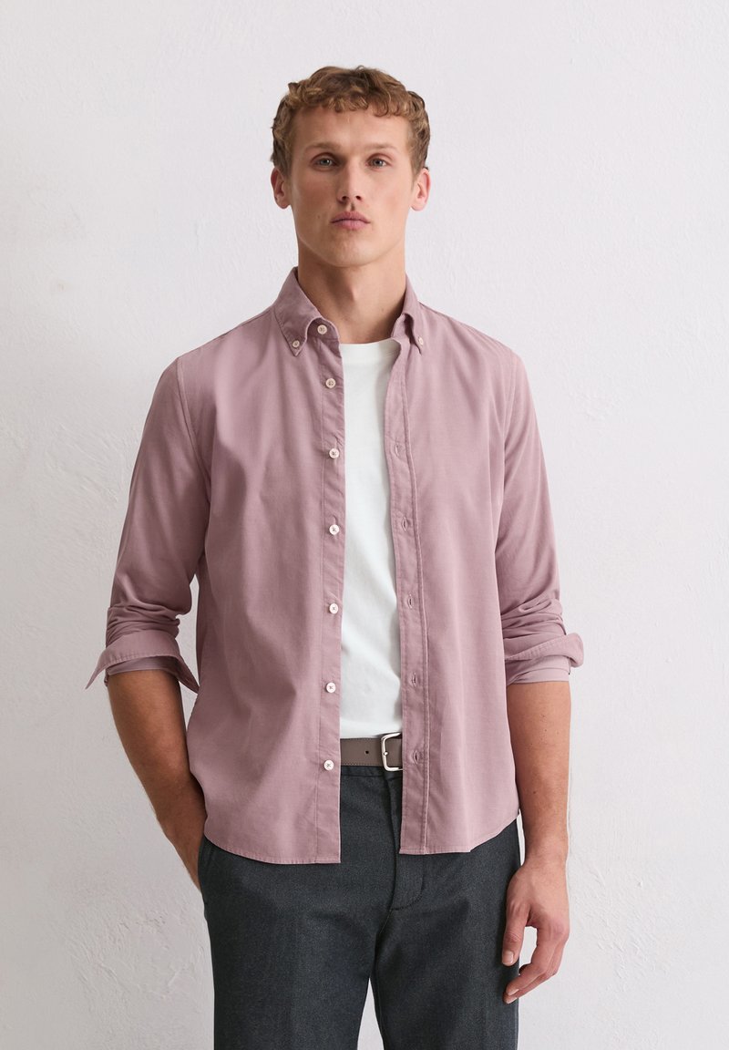 Jasnoróżowa koszula zapinana na guziki w luźnym kroju, z podwiniętymi rękawami, w zestawie z białym t-shirtem z okrągłym dekoltem i ciemnymi spodniami. Gładka tekstura materiału.