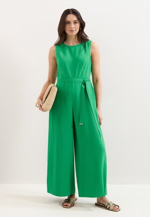 Vrouw die een mouwloze groene jumpsuit met wijde pijpen draagt, een gevlochten clutch vasthoudt en gouden sandalen en hoepeloorbellen draagt.