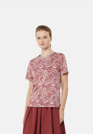 Jeune femme à la peau claire et aux cheveux blonds porte un haut rouge à manches courtes à motifs et une jupe plissée rouge unie sur fond blanc.