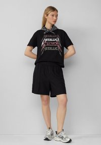 Zwarte graphic t-shirt met "METALLICA" in vette letters, gecombineerd met zwarte shorts en zilveren sportschoenen. Achtergrond is effen grijs.