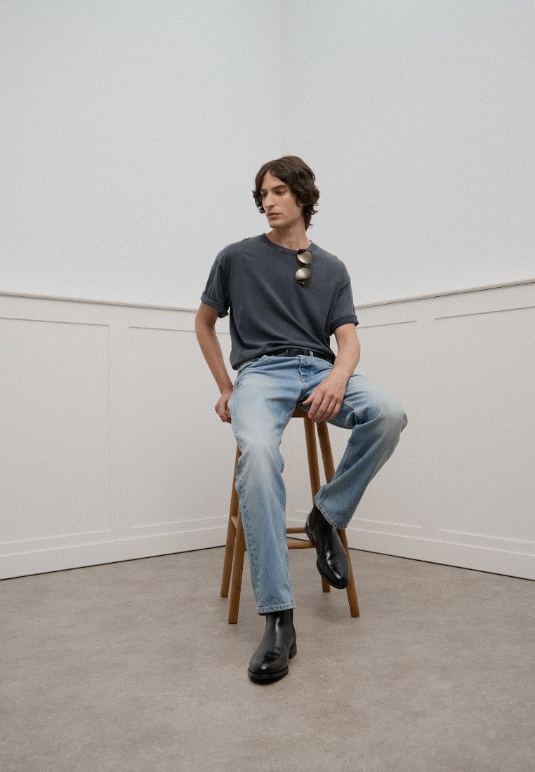 Graues kurzärmliges T-Shirt, hellblaue Jeans und schwarze Lederstiefel. Das Modell sitzt auf einem Holzstuhl in einem minimalistischen Innenraum.
