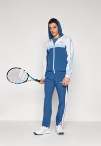 Blå joggedrakt med hette som har lyseblå og hvite paneler, sammen med hvite tennissko og holder en tenniskølle.