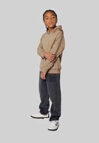 Sweatshirt à capuche beige avec un logo DKNY en relief, associé à un jean gris foncé comportant un imprimé de logo subtil, et des baskets de sport blanches.