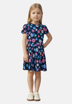 Katoenen kinderkleed met korte mouwen, marineblauwe achtergrond, bloemendruk in roze, blauw en wit, gelaagde ruches aan de onderkant, platte witte schoenen.