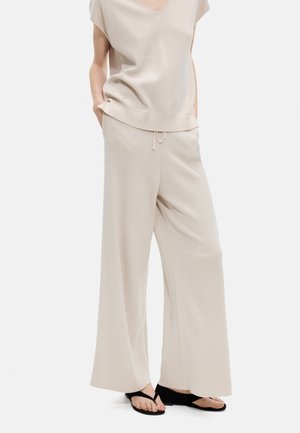 Osoba nosząca luźne beżowe dziergane spodnie i pasujący krótki top z dekoltem w serek, z rękami w kieszeniach, w czarnych sandałach na koturnie.