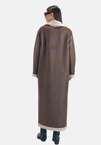 Bruine lange jas met een gestructureerde afwerking, voorzien van een crème shearling kraag en manchetten. Draagt over zwarte enkellaarsjes.