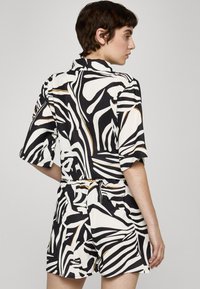 Combinaison courte à rayures noires et blanches avec col, manches courtes et taille cintrée ornée d'accents dorés. Texture de tissu lisse.