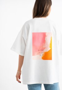 Witte oversized T-shirt met een grote abstracte print in tinten van roze, oranje en geel met tekst. Korte mouwen en ronde halslijn.