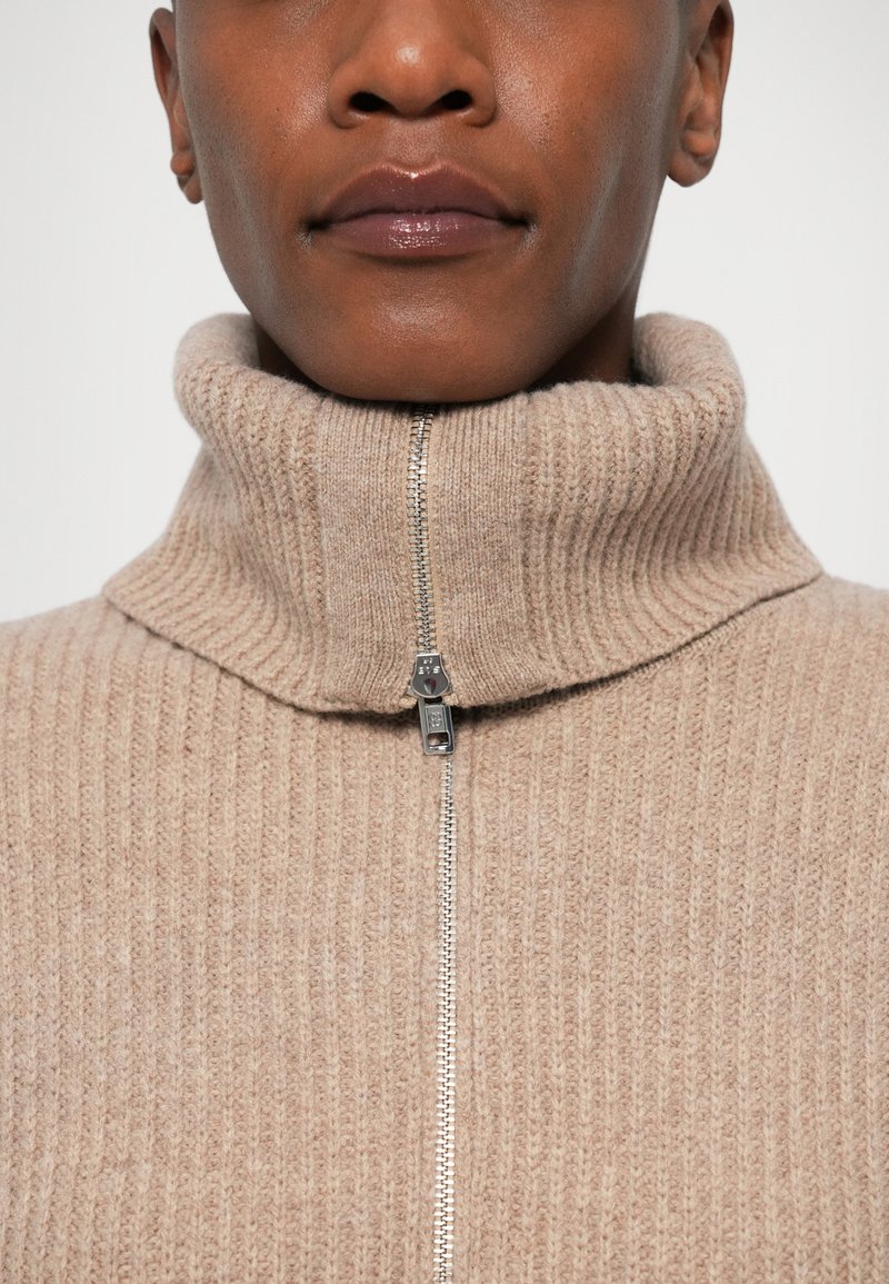 Närbild av en person som bär en beige ribbad stickad turtlenecktröja med en silverfärgad dragkedja som är delvis zipad.