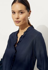 Marineblouse met een v-halslijn met ruffles, lange mouwen en een gladde, zachte textuur. De stof lijkt licht en soepel.