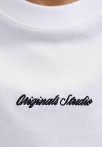 Tricou alb din bumbac, cu text brodat negru "Originals Studio" pe față. Textură netedă și design clasic cu guler rotund.