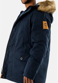 Giacca parka di colore blu navy realizzata in tessuto spesso e texturizzato, dotata di cappuccio foderato in pelliccia, tasche frontali e una patch in pelle con logo sul maniche.